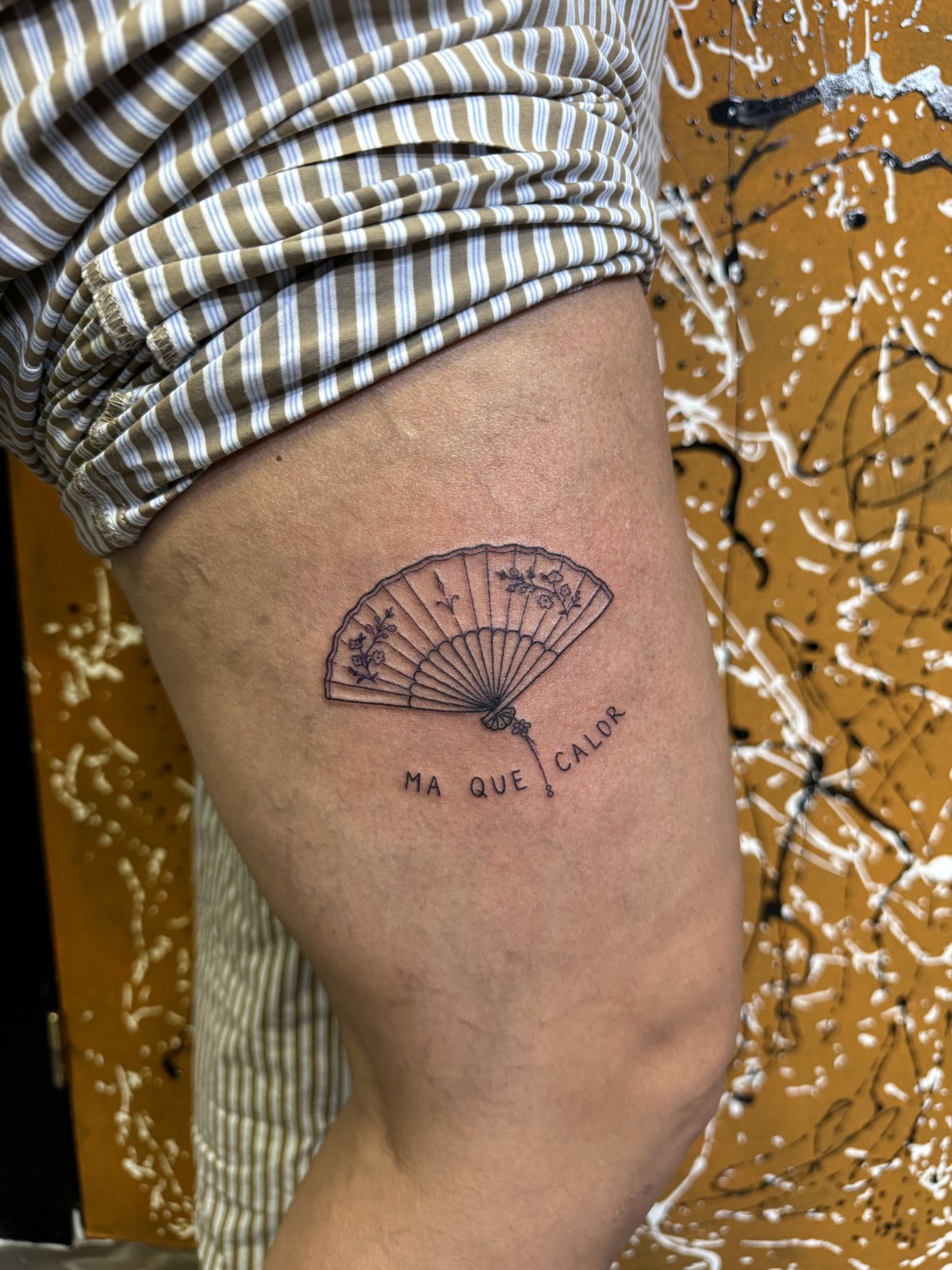 Fine line tattoo of a fan with flowers and lettering: ma que calor